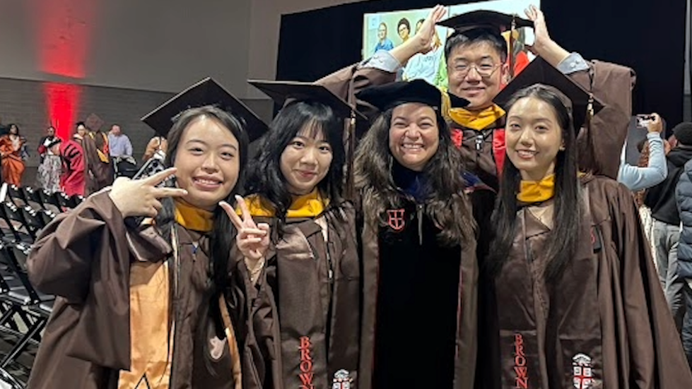 Five smiling graduates and faculty in Brown University academic regalia pose playfully together at an indoor commencement ceremony.